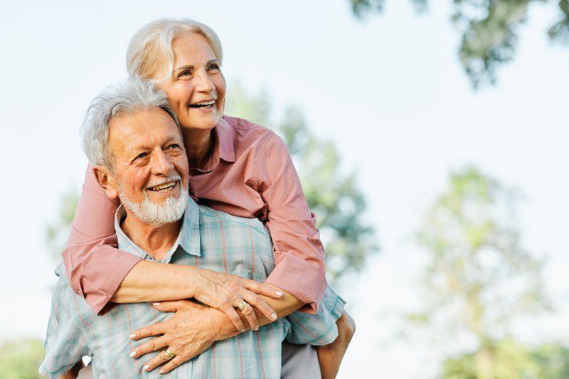 An older woman smiles while giving her partner a piggyback ride outdoors, surrounded by trees and greenery—a joyful moment celebrating couples and the vibrant community at Assured Assisted Living.