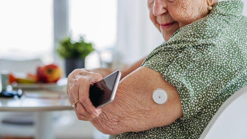 An older woman scans a white, dementia-friendly glucose monitor on her upper arm with a smartphone while sitting at a table.