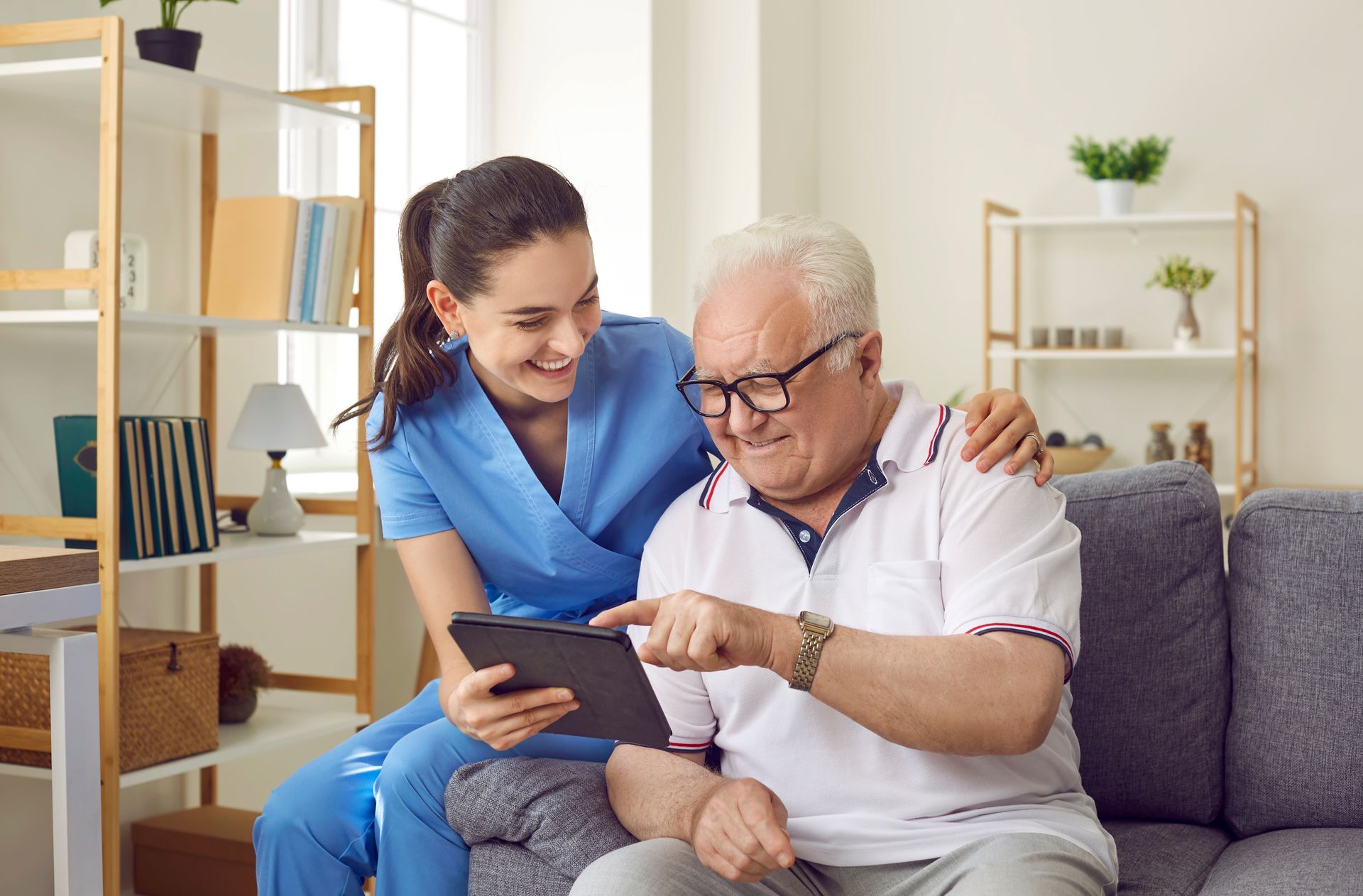 A nurse in blue scrubs sits beside an older man on a couch, both smiling and looking at a tablet as they review the latest research on TBI recovery together.