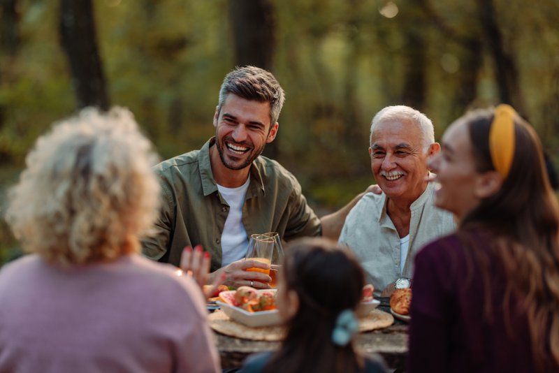 Four adults and one child sit around an outdoor table, sharing food and drinks, smiling and engaging in conversation with loved ones in a wooded setting.