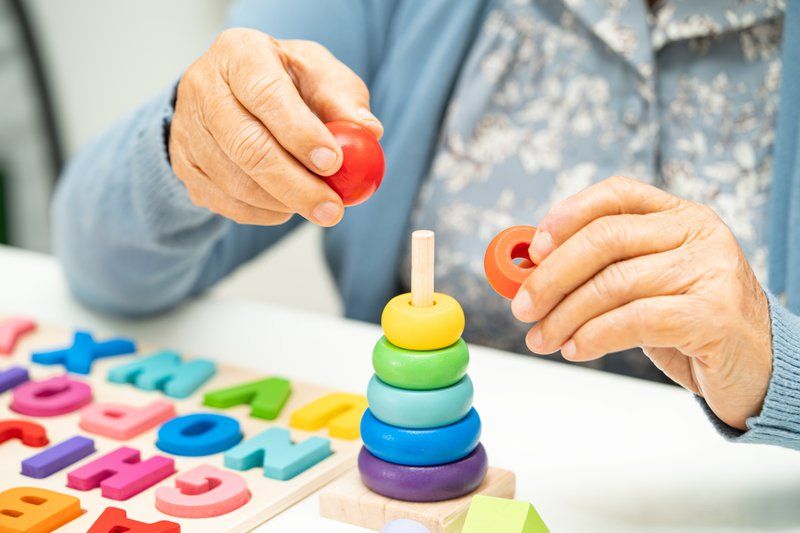 Close-up of an elderly person’s hands stacking colorful rings on a wooden toy, with foam alphabet letters on the table—highlighting potential for cognitive rehabilitation through engaging activities.