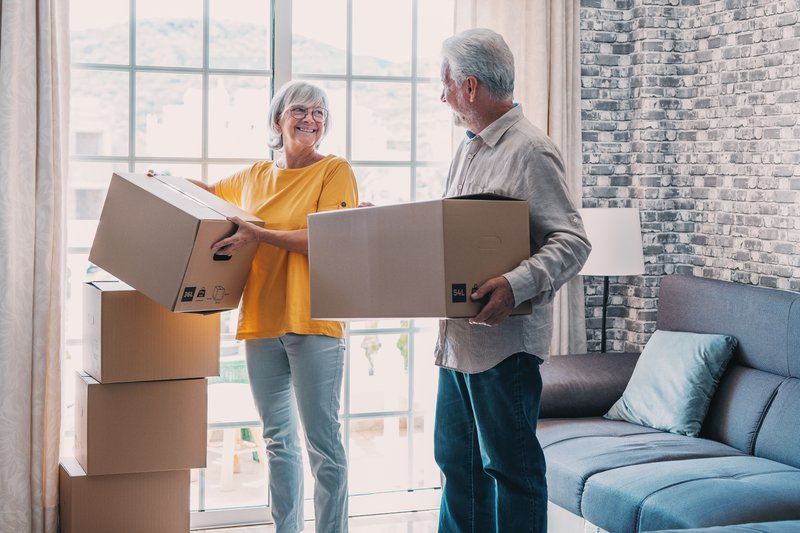 Two older adults stand in a living room holding cardboard boxes, with more boxes stacked nearby, suggesting a stress-free move or unpacking—perhaps following a helpful moving checklist for assisted living.