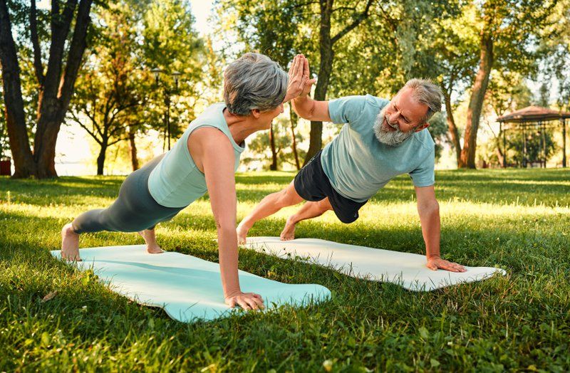 Two older adults exercise on yoga mats outdoors, holding plank positions and giving each other a high-five—a fun way to incorporate memory exercises and dementia activities into their active park routine.