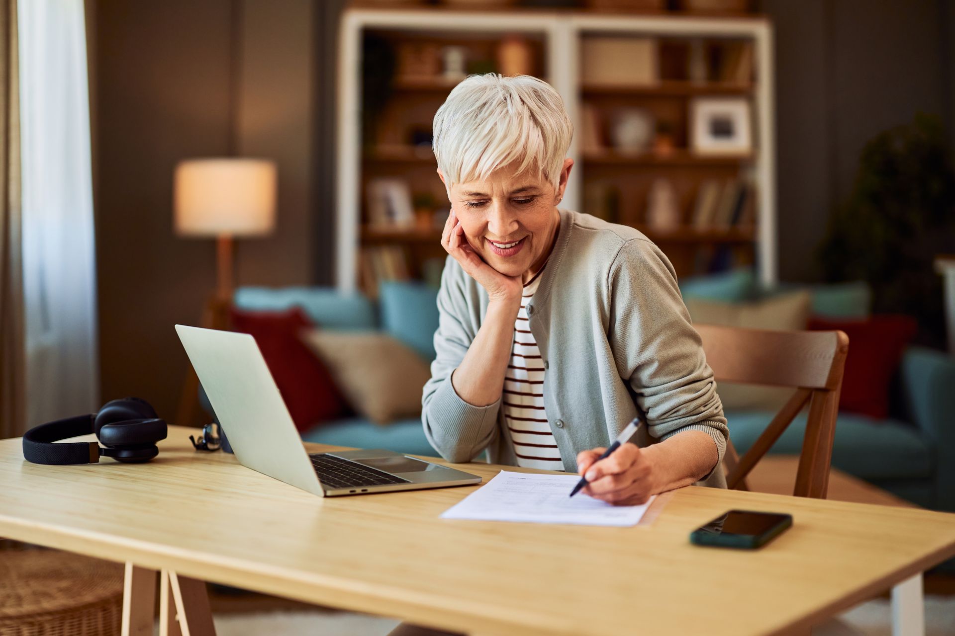 Smiling person with short gray hair writes about Advance Care Planning on paper at a desk with a laptop, headphones, and phone, in a cozy living room setting.