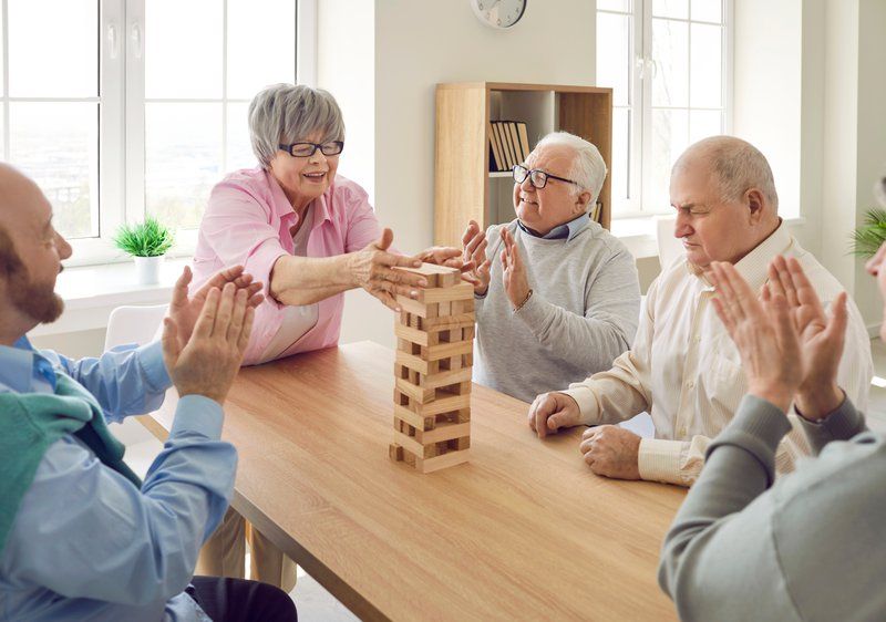 Five older adults sit around a table playing Jenga, with one woman pulling a block while others watch and clap in a bright, sunlit room, fostering understanding of behavioral changes often seen in dementia patients.