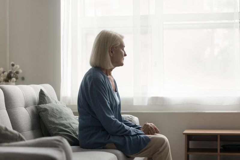 An older woman with gray hair sits on a sofa, looking out a bright window in a minimally furnished living room, quietly navigating grief as she finds moments of peace.