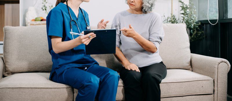 A nurse in blue scrubs talks with an older woman on a beige sofa, holding a clipboard and pen—demonstrating the attentive care found in quality assisted living facilities.