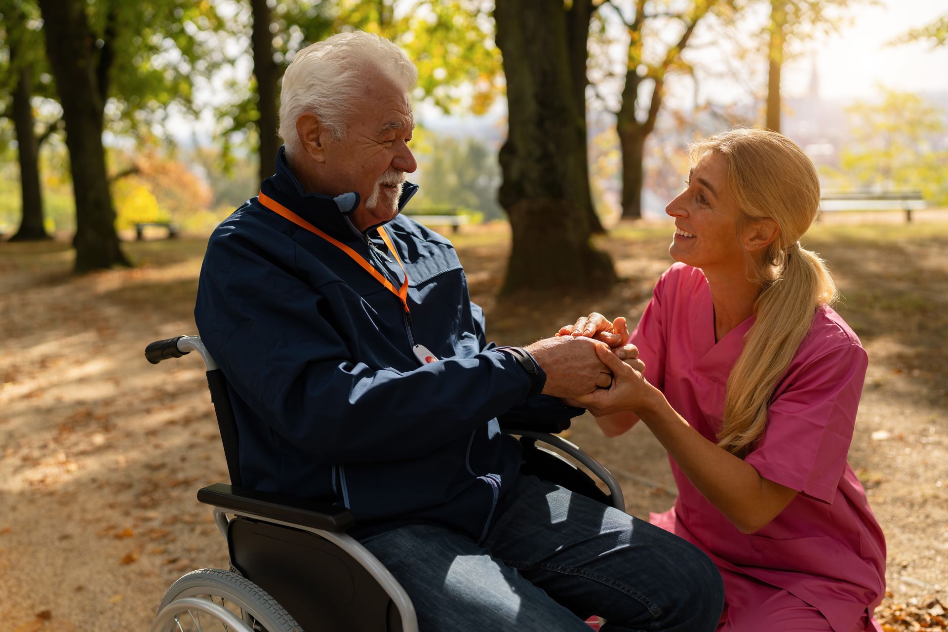 An elderly man in a wheelchair holds hands and smiles with a caregiver in pink scrubs while enjoying a sunlit park, highlighting the warmth and connection that complement dementia care technology.