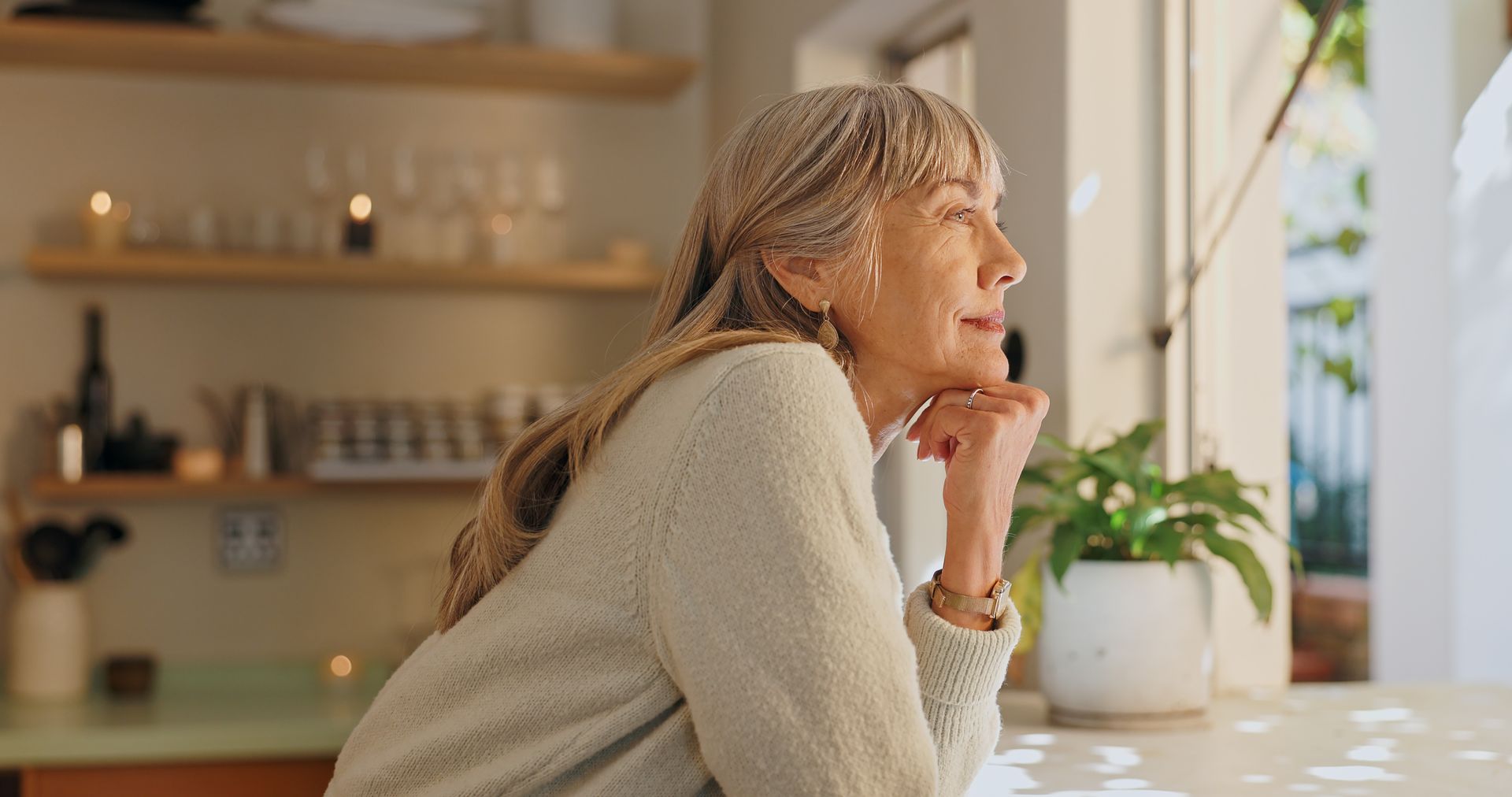 An older woman with long gray hair sits indoors by a window, resting her chin on her hand and looking thoughtfully outside, reflecting on loneliness. A potted plant is on the table beside her.