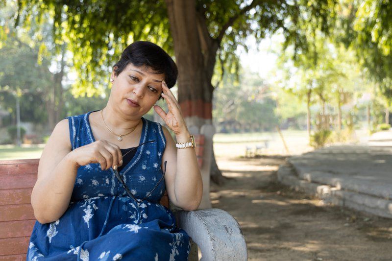 A woman in a blue dress sits on a bench outdoors, holding her glasses and resting her head on her hand, appearing thoughtful or stressed—possibly reflecting on early signs of dementia.