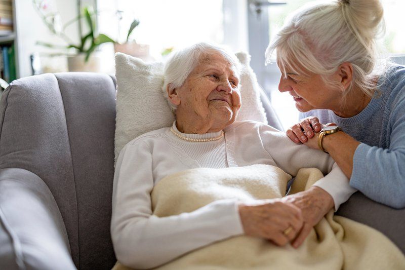 An elderly woman lies on a couch under a blanket while a middle-aged woman—perhaps engaging in volunteerism—leans in, smiling and holding her hand in the bright, homey room typical of warm senior communities.