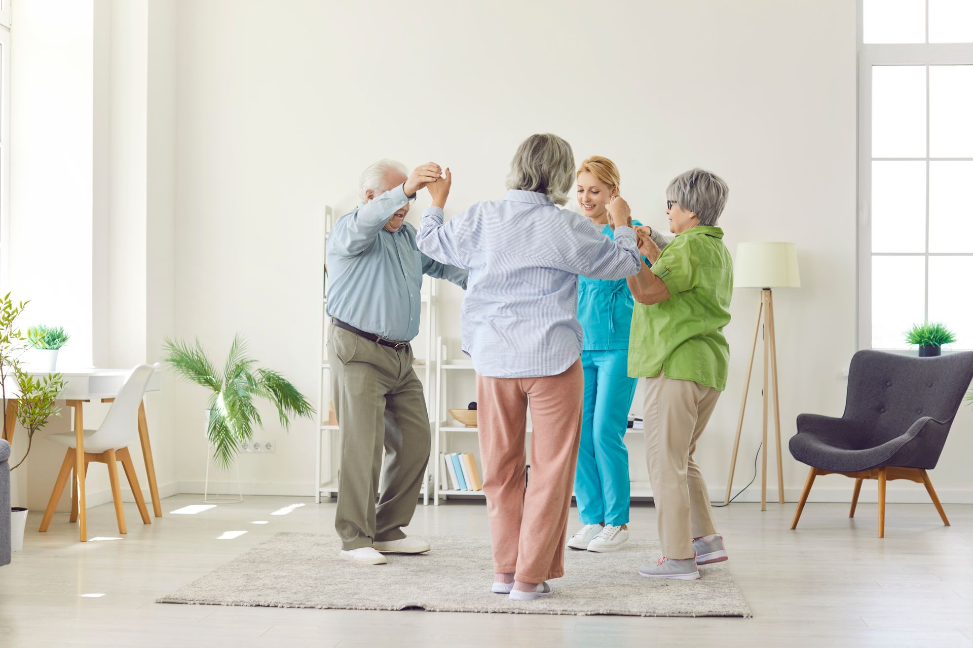 Four older adults and a caregiver stand in a circle holding hands and dancing in a bright, modern assisted living facility.