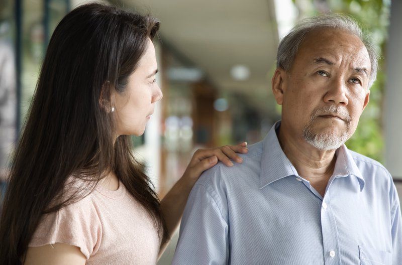A woman places her hand on an older man's shoulder, offering comfort and understanding. The man looks away with a serious expression—capturing the effects on families living with Alzheimer’s Disease. They are outdoors, blurred buildings in the background.