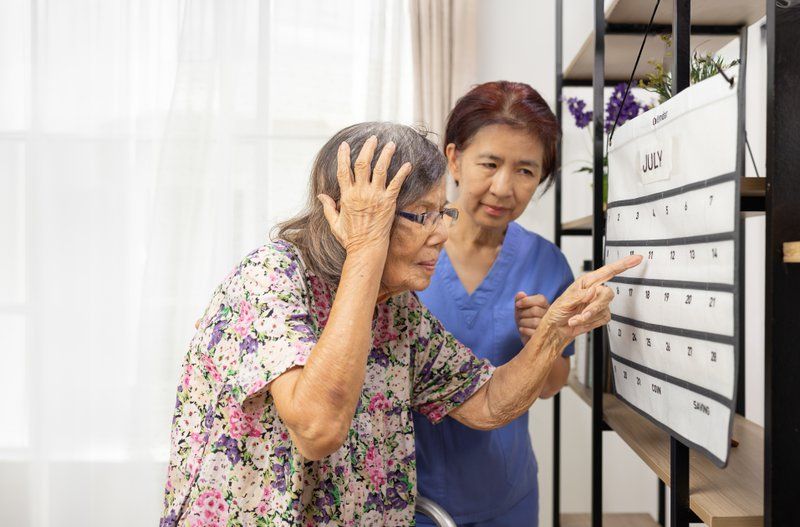 An elderly woman looks confused while pointing at a wall calendar, with a caregiver beside her in a bright room—a scene often observed among dementia patients experiencing behavioral changes.