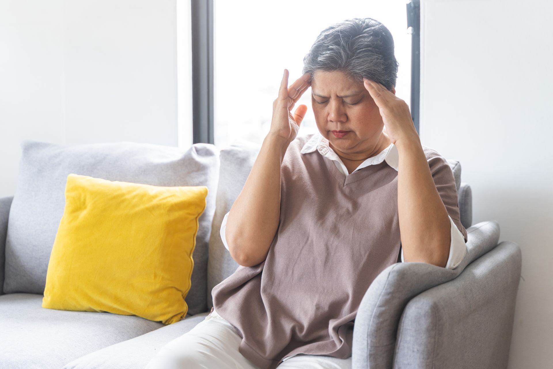 An older woman sits on a couch with her hands on her temples and eyes closed, appearing to have a headache or discomfort, possibly experiencing post-injury seizures.