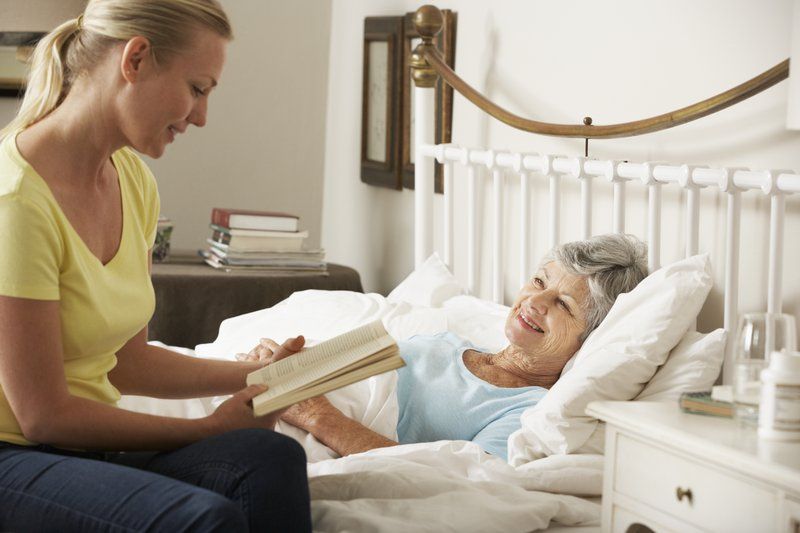 A woman sits beside an elderly woman lying in bed, reading a book to her in a bright bedroom, offering gentle palliative care.
