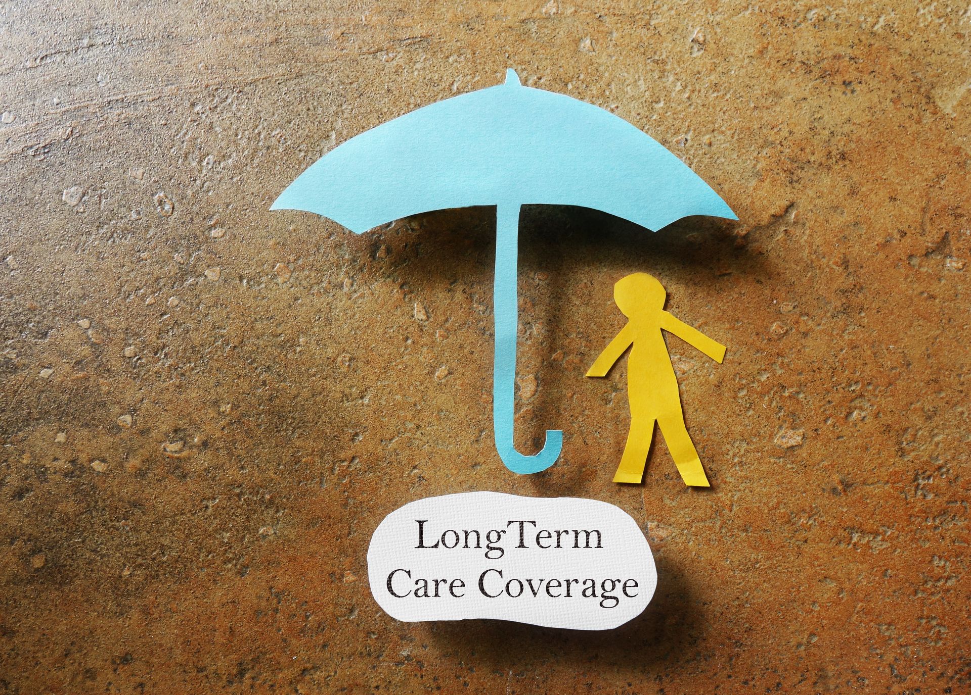 Paper cutouts of a blue umbrella and a yellow person sit beside a label reading "Long Term Care Coverage," symbolizing the protection offered by long-term care policies on a textured brown surface.