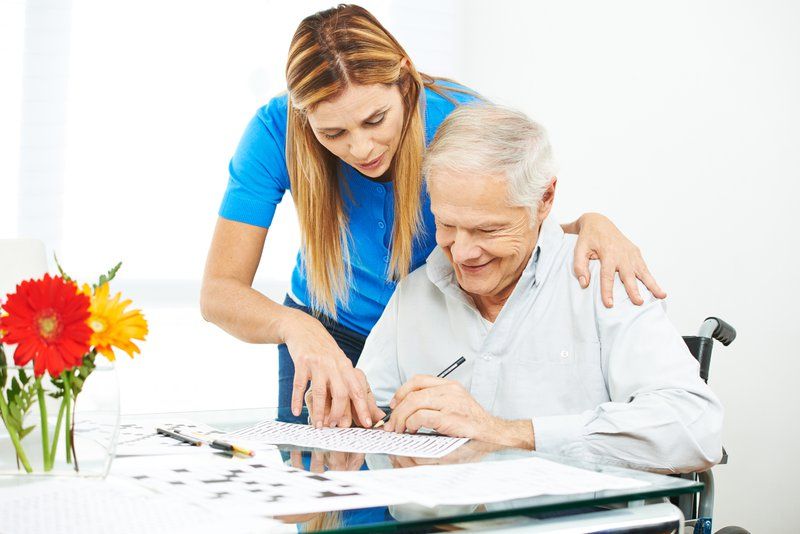 A caregiver provides personalized care as she assists an elderly man in a wheelchair with a crossword puzzle at a table, offering support for dementia treatment. A vase of flowers adds warmth to the scene.