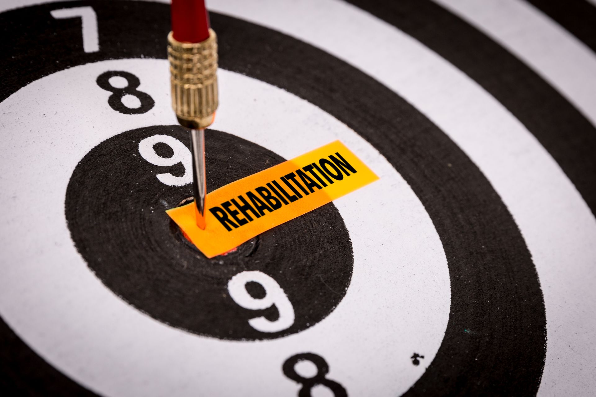 A dart hits the bullseye of a black and white target, piercing a yellow strip labeled "REHABILITATION," symbolizing the precision needed in setting rehabilitation goals along your rehabilitation journey.