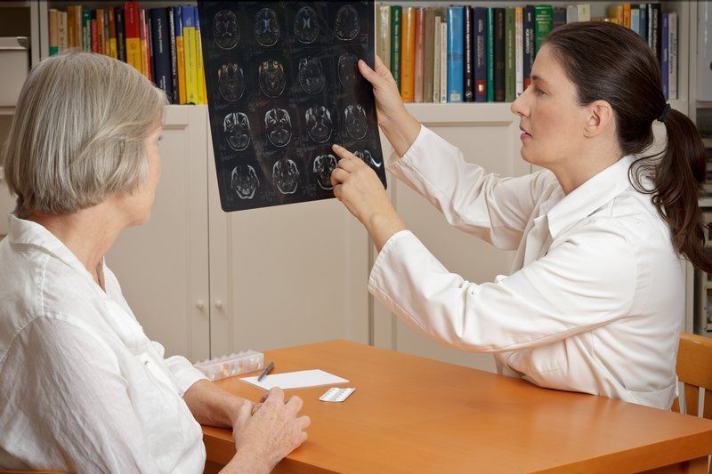 A doctor shows and explains brain scan images to an older woman seated at a desk in a medical office, discussing brain injury basics and the potential impact on loved ones.