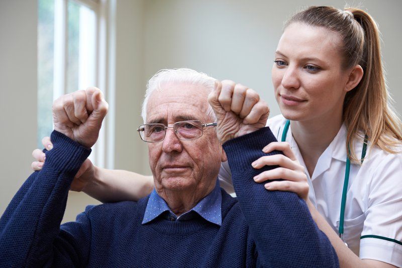 An older man sits indoors with his arms raised while a healthcare professional assists him during a physical therapy session, supporting his mobility and well-being through the various dementia stages.
