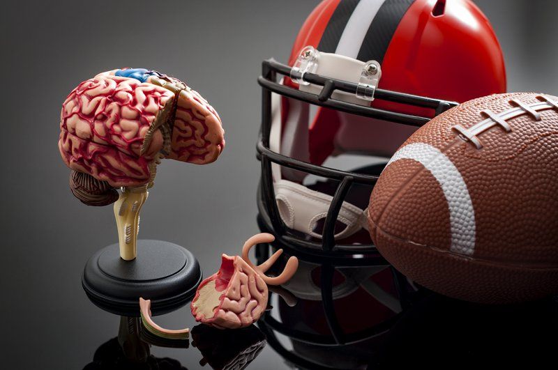 A model of a human brain with a detachable section is displayed next to an American football helmet and a football on a reflective surface, highlighting the link between sports and Traumatic Brain Injury.