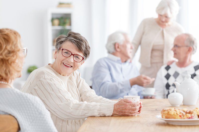 A group of elderly people sits at a table in a bright, cozy room, enjoying tea, pastries, and lively conversation—highlighting the importance of social engagement in memory care communities.