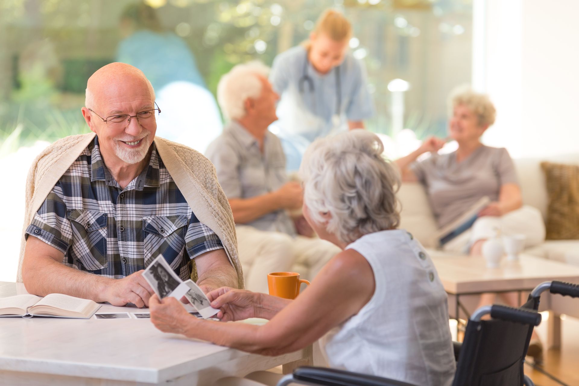 Elderly man and woman sit at a table looking at photos, while other seniors and a caregiver engage in the bright common room, reflecting the warm community of Assured Senior Living and its focus on changing health needs in senior care.