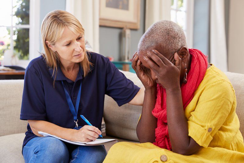 A woman in a yellow dress sits on a couch with her head in her hands while another woman next to her takes notes and offers support, highlighting the importance of supportive care plans for those living with dementia.
