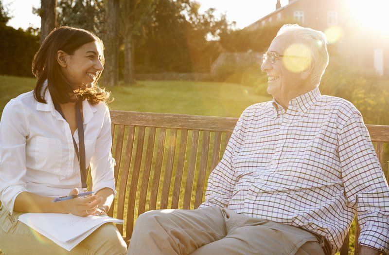 A young woman with a clipboard sits on a bench, smiling and talking with an older man in glasses and a checkered shirt, outdoors in sunlight—highlighting the importance of caregiver support for family caregivers.