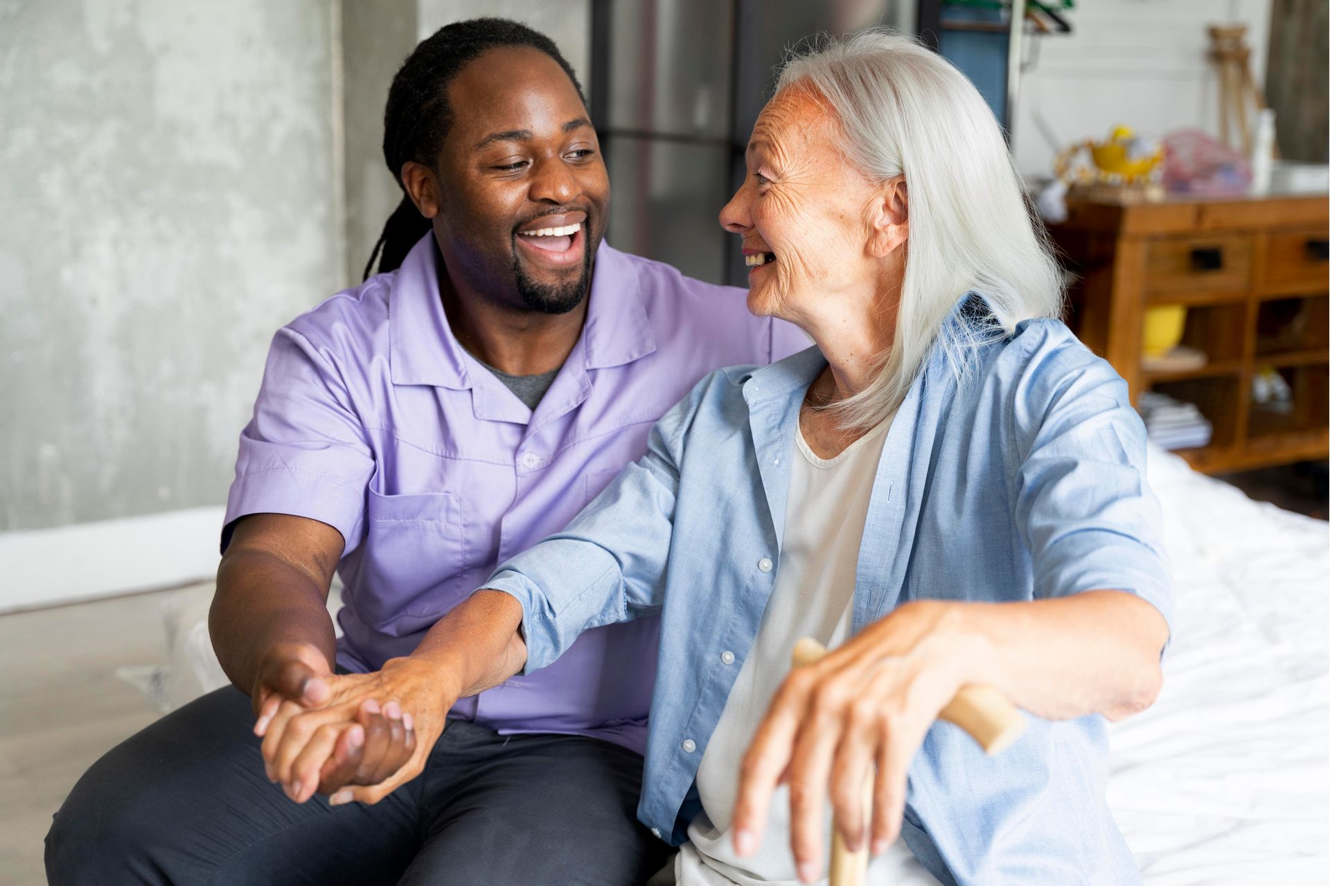 A young man and an older woman sit together indoors, smiling and holding hands. The woman holds a cane, and both appear to be enjoying a pleasant conversation—highlighting the importance of family caregivers for seniors.