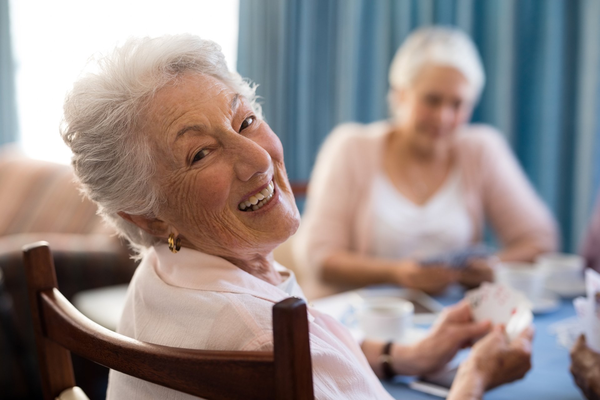 Elderly woman smiling at the camera while playing cards at a table, enjoying social activities that make transitioning to assisted living more enjoyable, with another woman in the background.