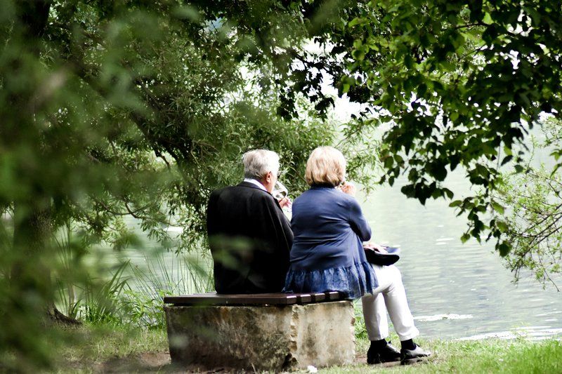 Two older adults sit on a bench by a lake, surrounded by green trees, enjoying the water as they maintain their independence in a safe, peaceful environment.